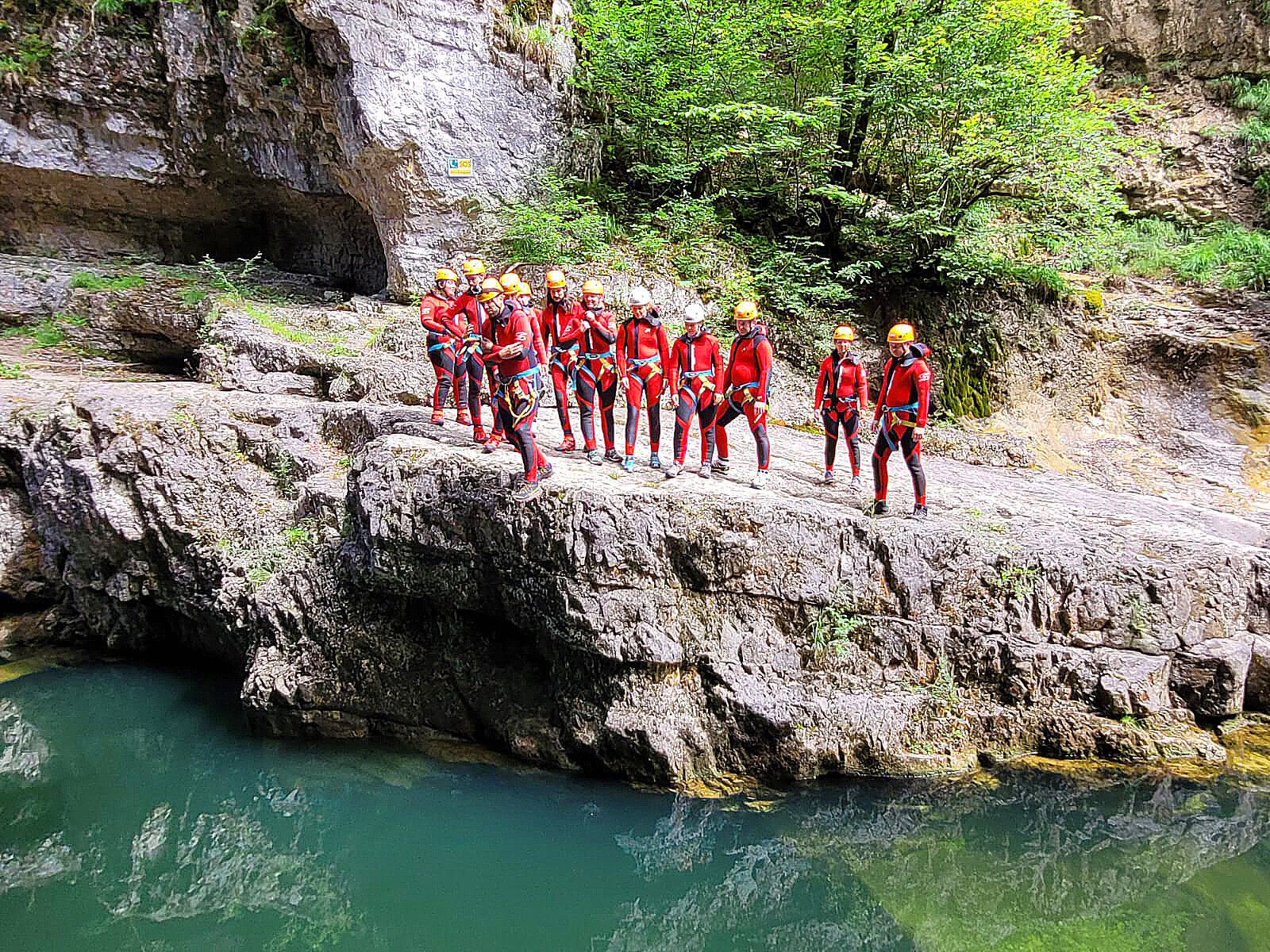 Gruppe in Neoprenanzügen und Helmen beim Canyoning, steht auf Felsvorsprung über türkisgrünem Wasser, bereit zum Sprung.