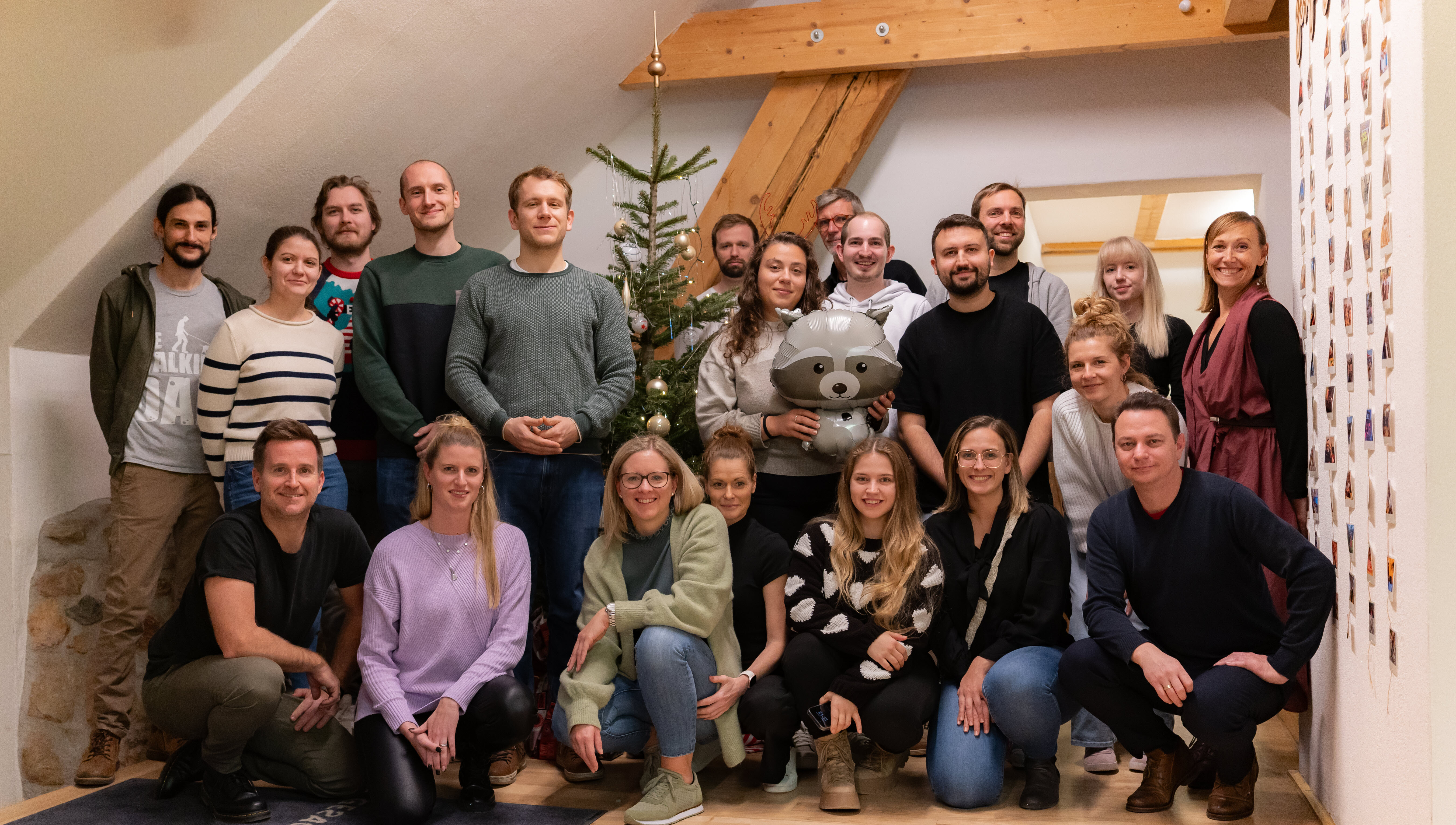 Teamfoto im Büro neben kleinem Weihnachtsbaum; lachende Gruppe, eine Person hält einen Waschbär-Ballon.