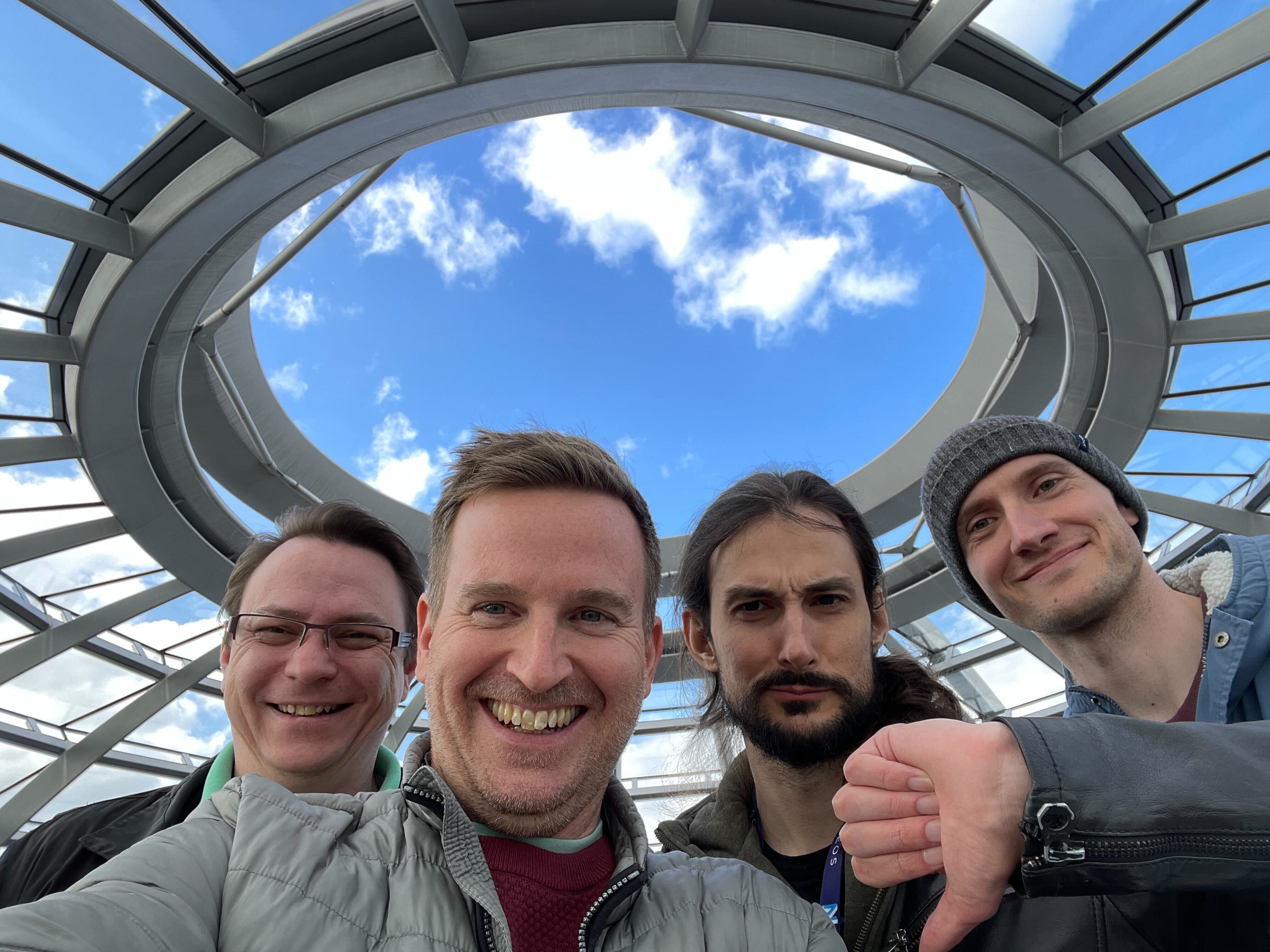Vier Personen machen ein Selfie unter einer modernen Glaskuppel mit blauem Himmel; drei lächeln, einer zeigt den Daumen nach unten.