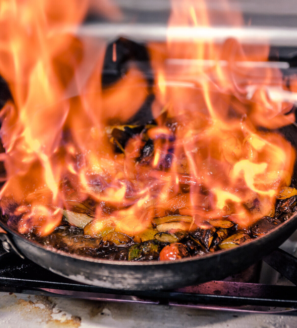 Flammen schlagen aus einer Pfanne beim Flambieren von Gemüse auf einem Gasherd in einer Profiküche.