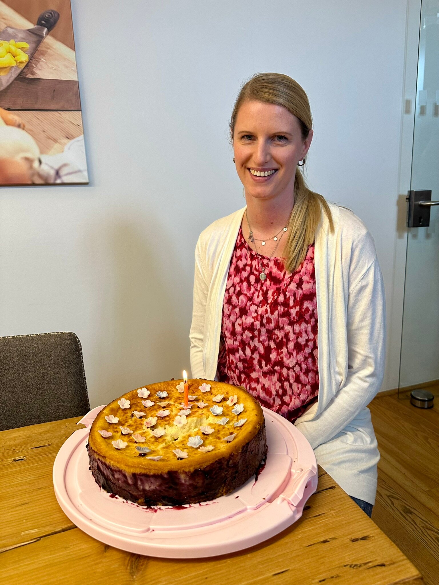 Lächelnde Frau am Tisch vor einer runden Torte mit Blütendeko und einer brennenden Kerze auf rosa Platte.