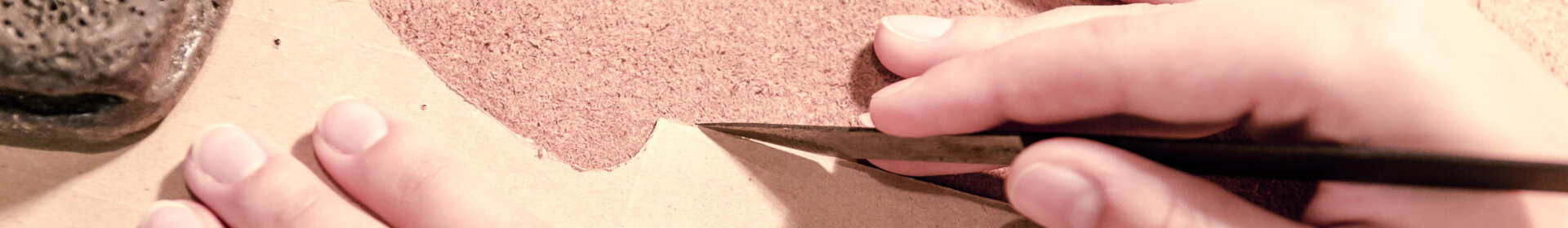 Zwei Hände schneiden mit einem Messer eine Lederform entlang einer Papierschablone.